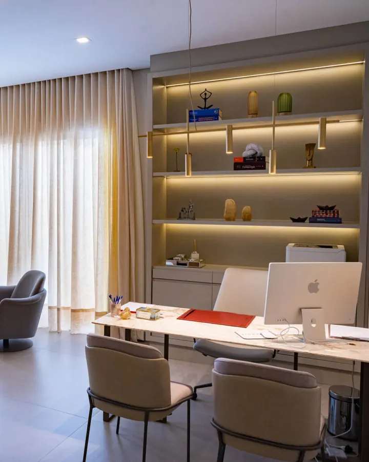 Consultório da Clínica Synergie com estante iluminada, cortinas claras e mesa de atendimento
